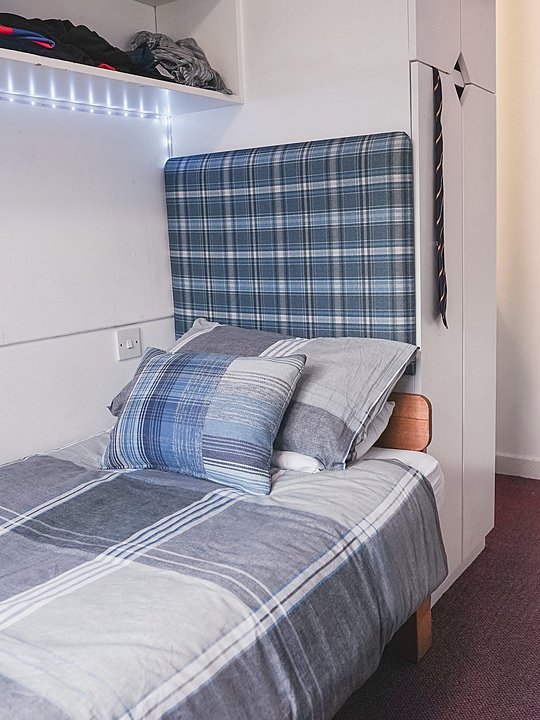 Neatly made boarding school bedroom with blue plaid bedding and built-in shelving.