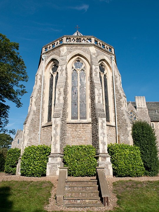 Historic stone chapel with tall arched stained-glass windows and decorative stonework, surrounded by greenery under a clear blue sky.