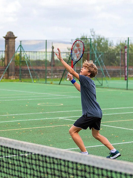 A student playing tennis outdoors, preparing to serve on a green court surrounded by fencing.