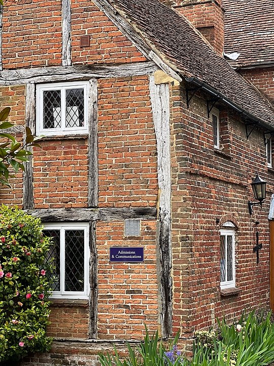 Old red-brick half-timbered building with leaded windows and a sign reading “Admissions & Communications,” surrounded by plants and flowers
