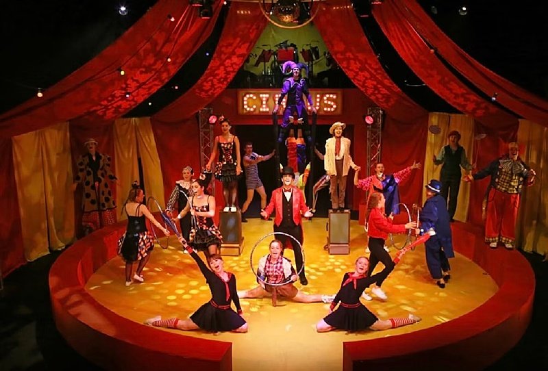 A group of performers in a circus-themed stage production with vibrant red curtains and spotlights.