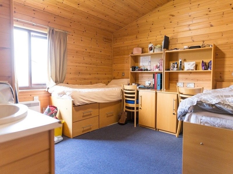 A cosy wooden dormitory room with two beds, desks, and bookshelves, featuring warm wooden paneling and a soft carpet.