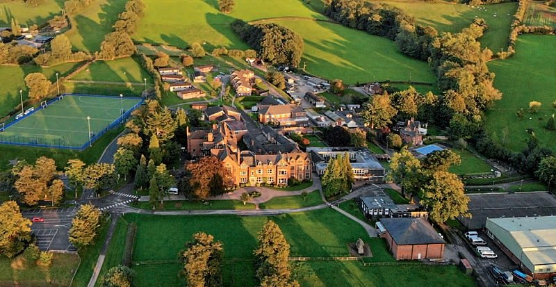An aerial view of a large countryside school campus surrounded by green fields, with multiple buildings, sports pitches, and tree-lined paths.