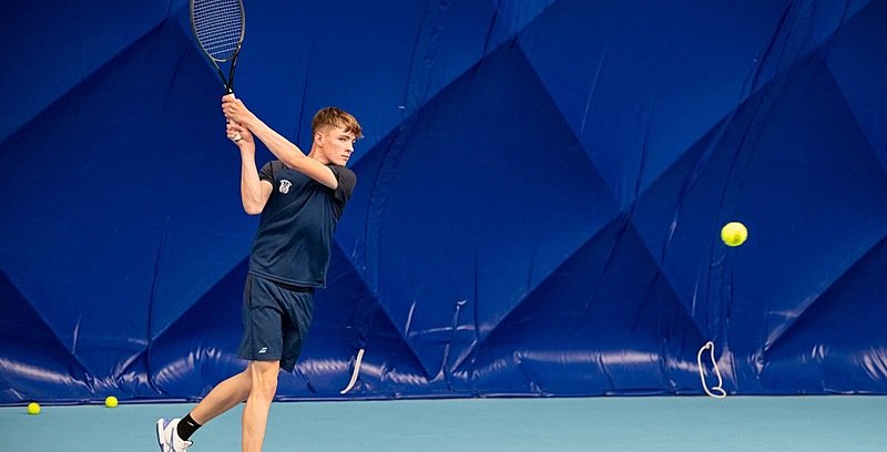 A boy with a Merchiston Castle tennis jersey swinging a tennis racket, ball flying towards him