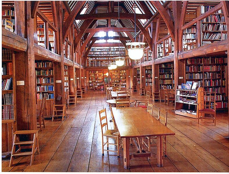 A spacious wooden library hall with high ceilings, rows of bookshelves, and long wooden tables for reading and study.