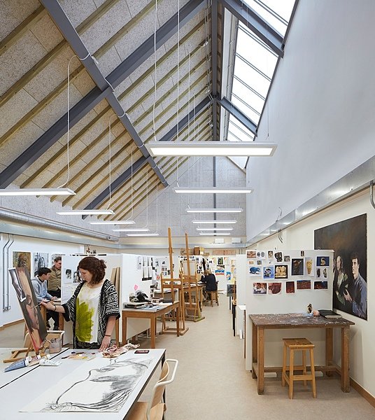 An art studio with tall skylights and rows of workstations where students paint and draw.