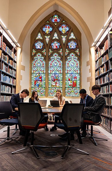 Students studying in a library with bookshelves and a large stained glass window behind them.
