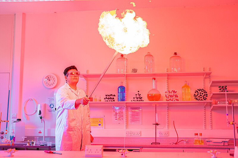 Student in a lab coat performing a flame experiment in a science classroom with pink lighting.