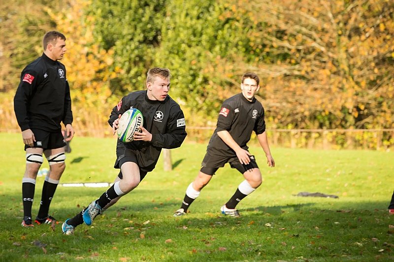 Pupils in black sports kits playing rugby on a grassy field with autumn trees in the background.