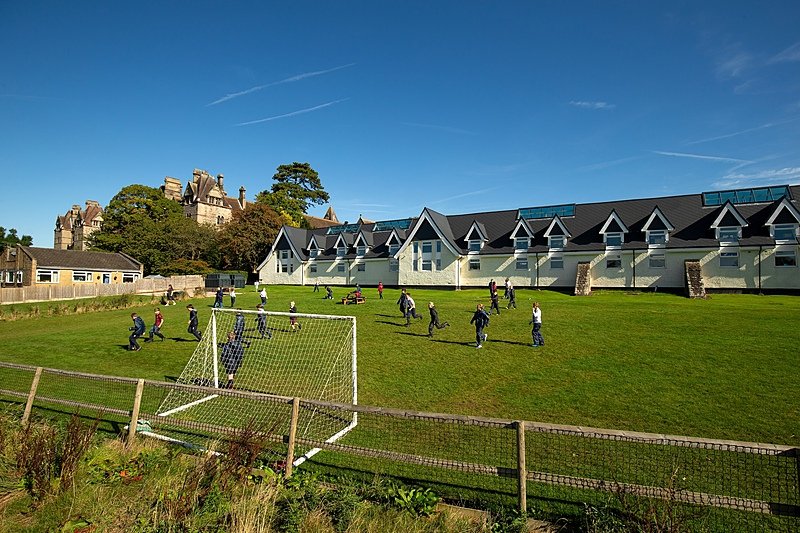 Pupils playing football on a grassy school field on a sunny day, with a modern building and an older, grand stone building in the background.