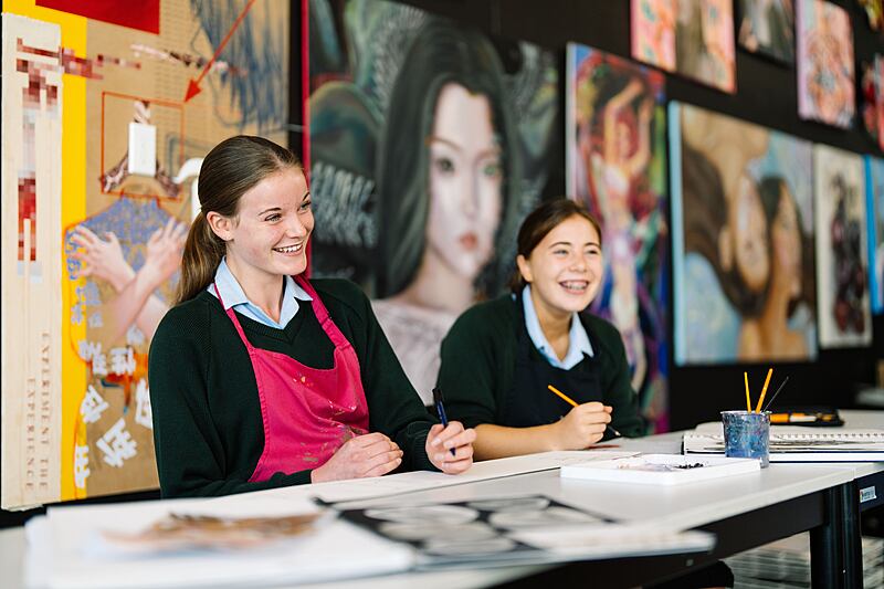 Two pupils sit at a table drawing, with large portrait artworks displayed on the wall behind them.