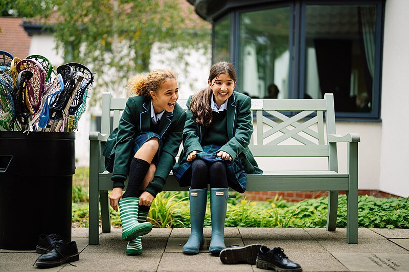 Zwei Schülerinnen in Schuluniform sitzen auf einer Holzbank vor einem Gebäude und sprechen miteinander.