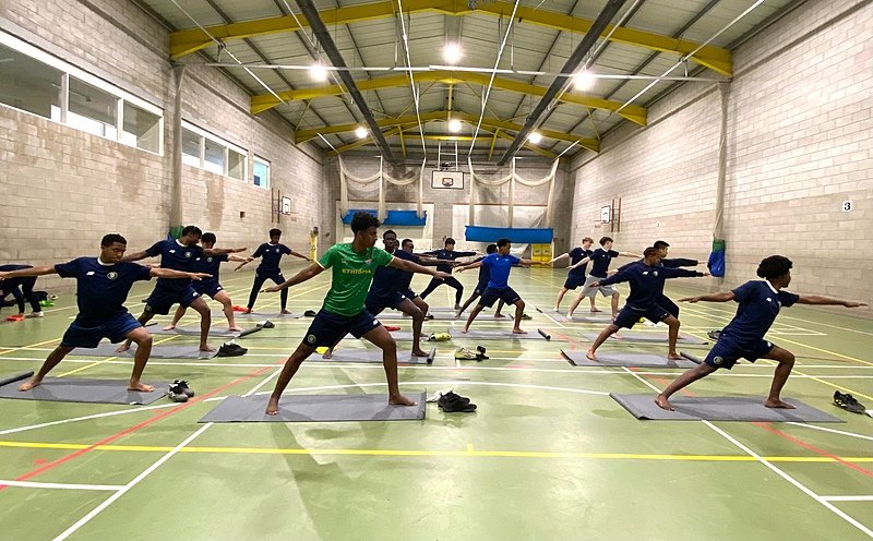 Students practise yoga poses together in a large indoor sports hall with high ceilings and bright lighting.