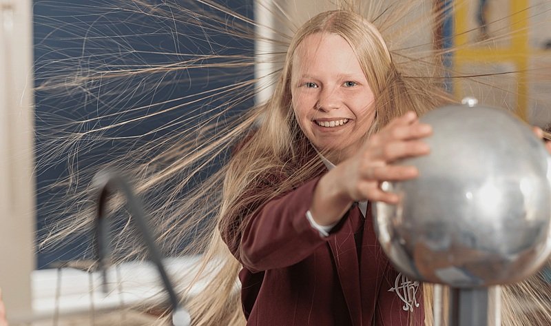 A smiling student in a science classroom touches a Van de Graaff generator as their hair stands on end from static electricity.