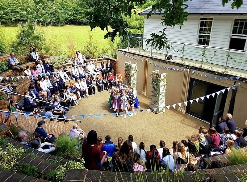 Students perform an outdoor play in a small amphitheatre surrounded by an audience seated on tiered brick steps decorated with bunting.