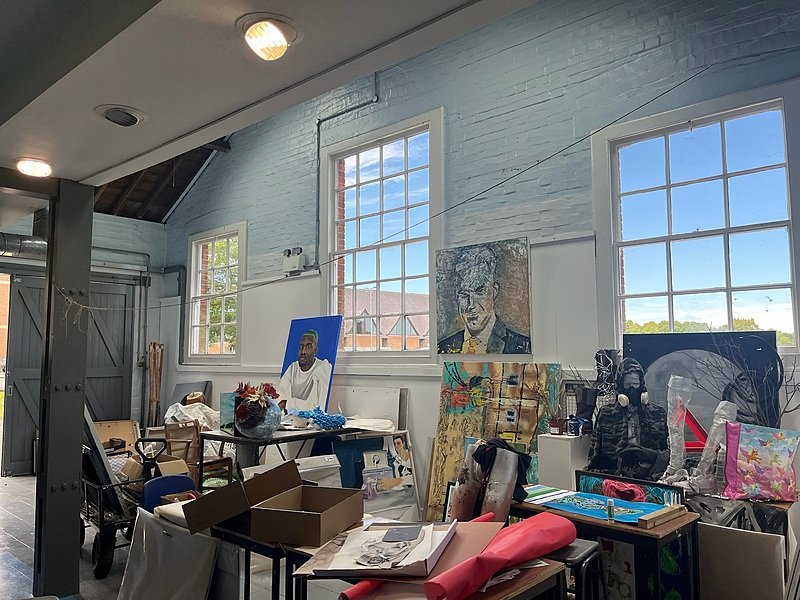 An art studio with large windows and bright natural light, showing various artworks including portraits and abstract paintings displayed around the room.