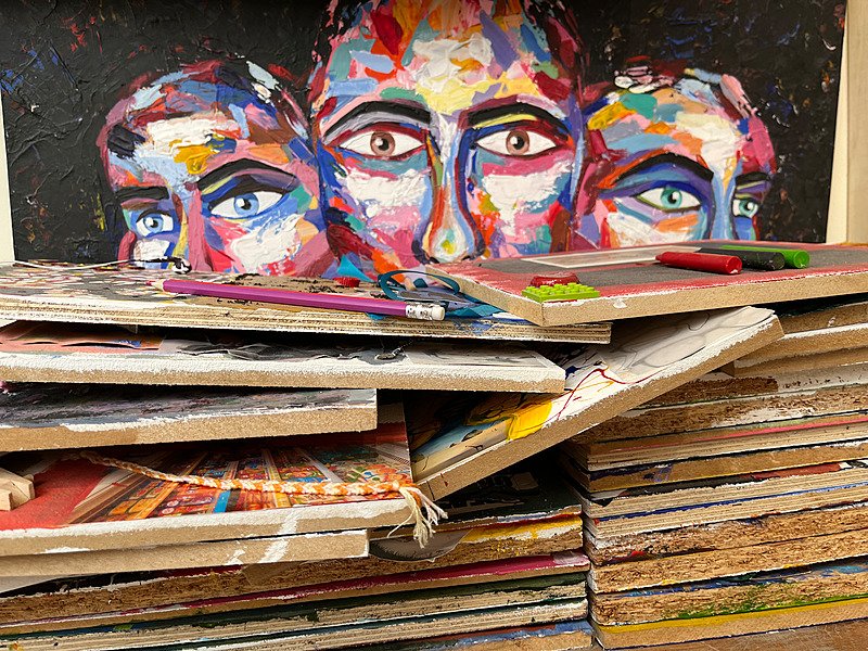 Several art canvasses on top of each other with a painting with three faces in the back