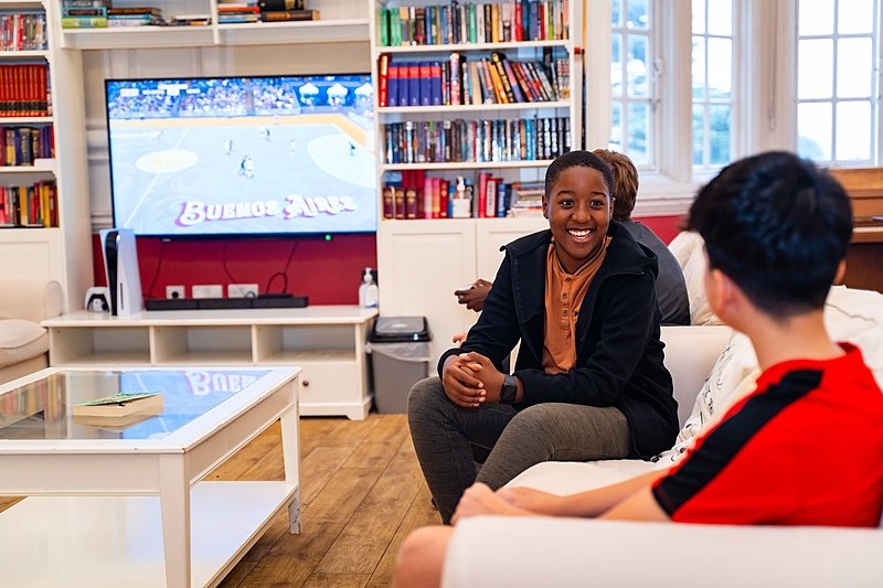 Boys are sitting in a boarding room, speaking and watching television.
