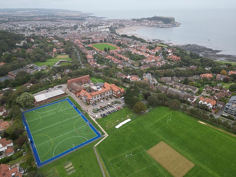 An aerial view shows a coastal school with sports fields, red-roofed buildings, and a view of the sea in the background.