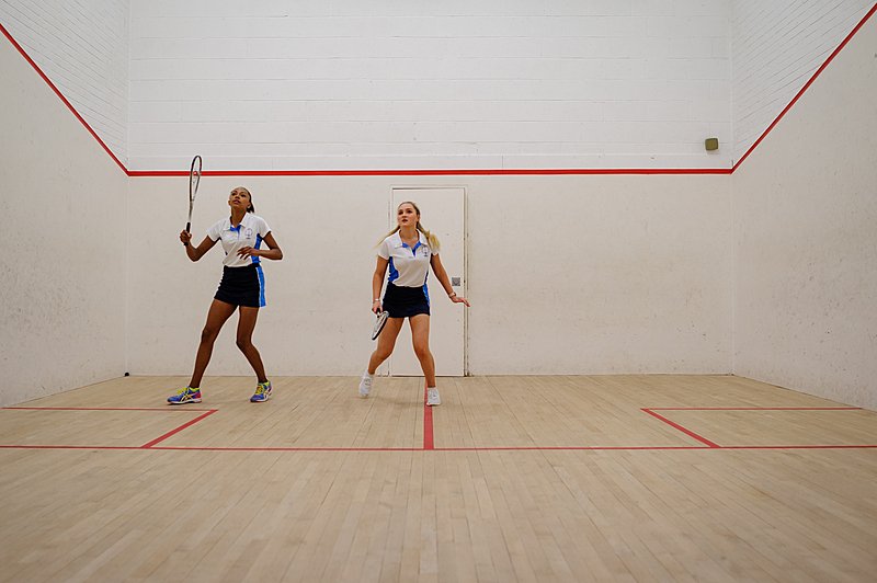 Two students play squash on an indoor court with wooden flooring and red boundary lines.
