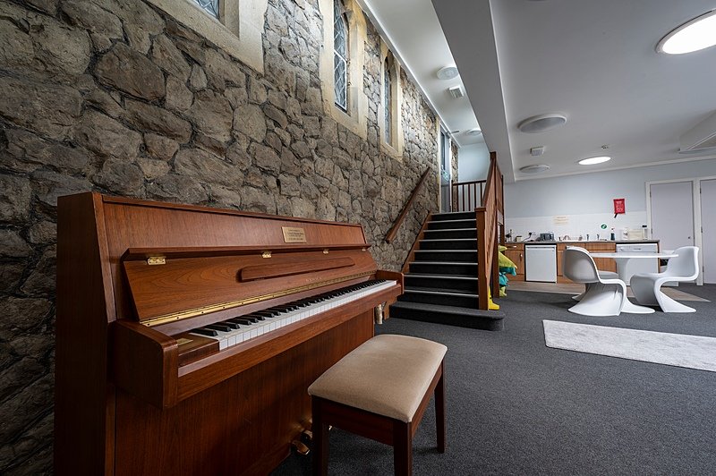 A cozy interior with a wooden piano and stool against a stone wall, next to a staircase leading up to a small kitchen and seating area with modern white chairs.