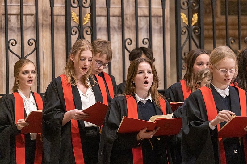 A group of students dressed in black robes with red stoles sing in a choir, holding red songbooks in front of a decorative wrought-iron backdrop.