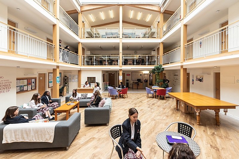 Spacious, modern boarding house atrium with several floors, wooden accents, sofas, and tables where pupils in school uniforms are sitting and talking.