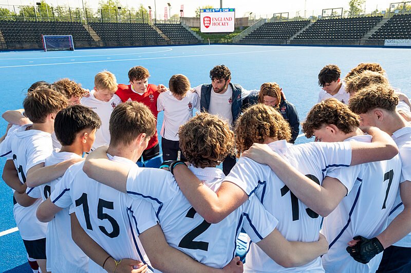 Boys’ hockey team in white jerseys standing in a circle with arms around each other on a blue hockey field, listening to their coach.
