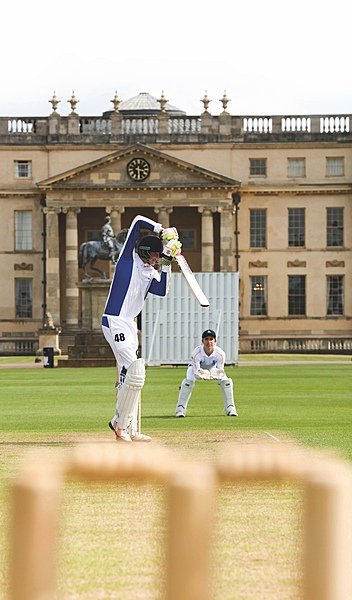 A batter swinging his bat on a cricket pitch with a wicketkeeper in position outside the school