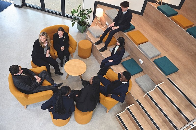 eight students in modern school uniforms sitting in a circle on armchairs, stools and stairs, seen from above