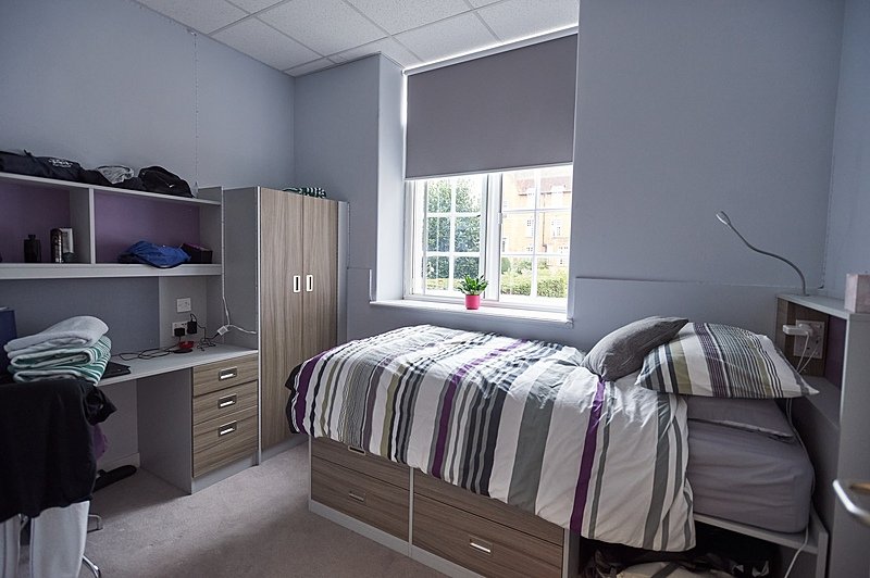 A tidy boarding school bedroom with striped bedding, a desk, and a wardrobe under a bright window.