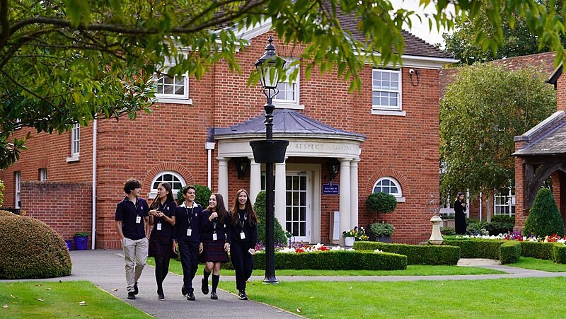 Group of pupils in school uniform walking and chatting in front of a red-brick school building surrounded by gardens.