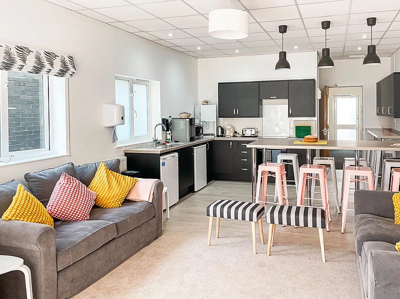 A bright and cosy community room with sofa and open-plan kitchen and dining in a boarding house