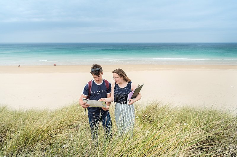 A boy and a girl in the dunes by a beach looking at a map
