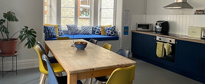 A cosy boarding house kitchen with a window bench with cushions and a big dining table with colorful chairs