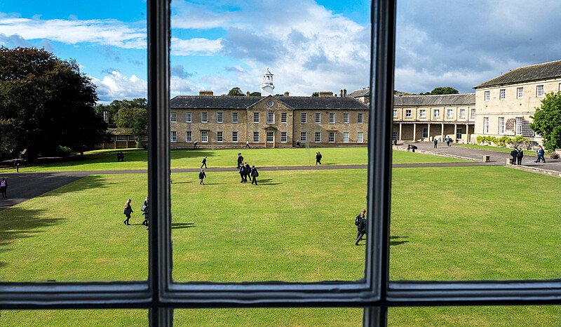 A view from a window overlooking a school courtyard where students walk across a large grassy area surrounded by stone buildings.